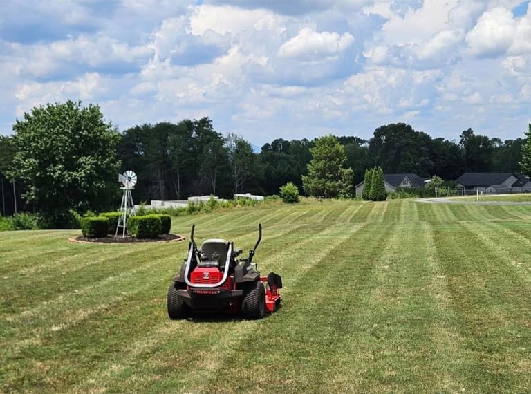 Red and black riding lawnmower mowing a striped grass field with trees and blue sky in background