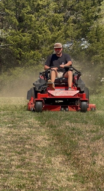 Man operating a red zero-turn riding lawnmower on a grass field with trees in the background