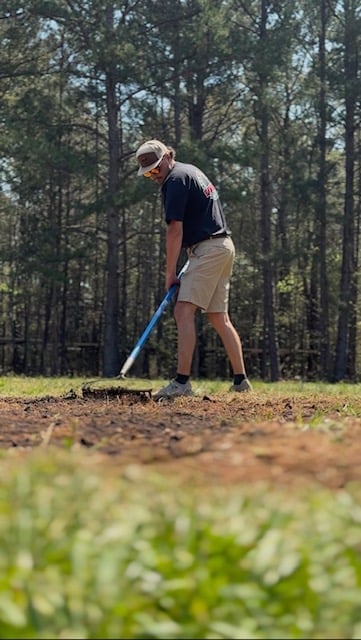 Man in dark shirt and khaki shorts using a blue shovel to dig in brown soil with forest trees in background