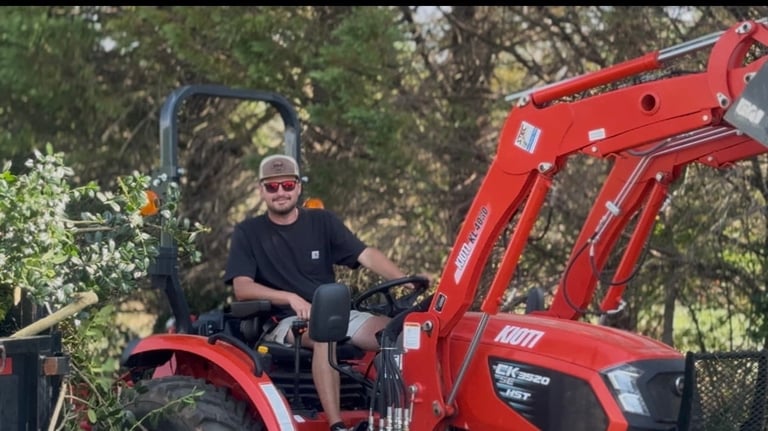 Man operating a red compact tractor with front-end loader in a wooded area